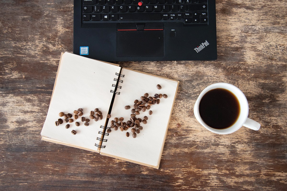 Looking a brown wooden table with cup of coffee, laptop and coffee beans poured over laptop