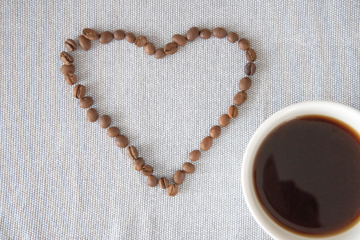 White cup with black coffee on a table, coffee beans arranged in heart shape next to it
